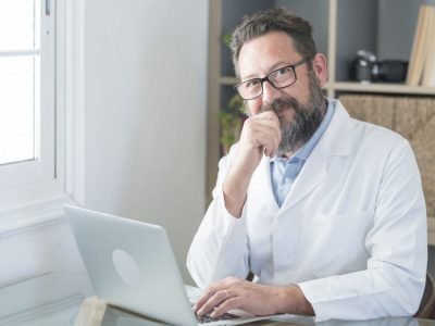 smiling-old-male-doctor-in-glasses-and-white-uniform-sit-at-desk-in-hospital-work-on-laptop.jpg